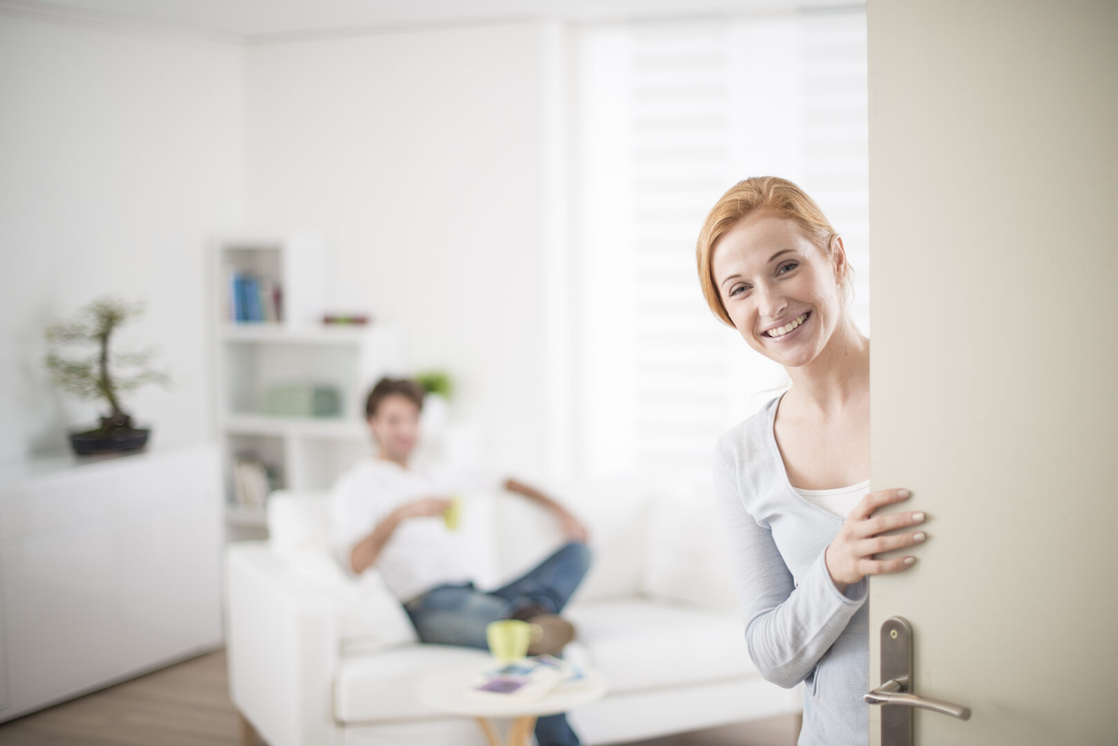 Woman smiling at apartment door with person relaxing in the room behind her