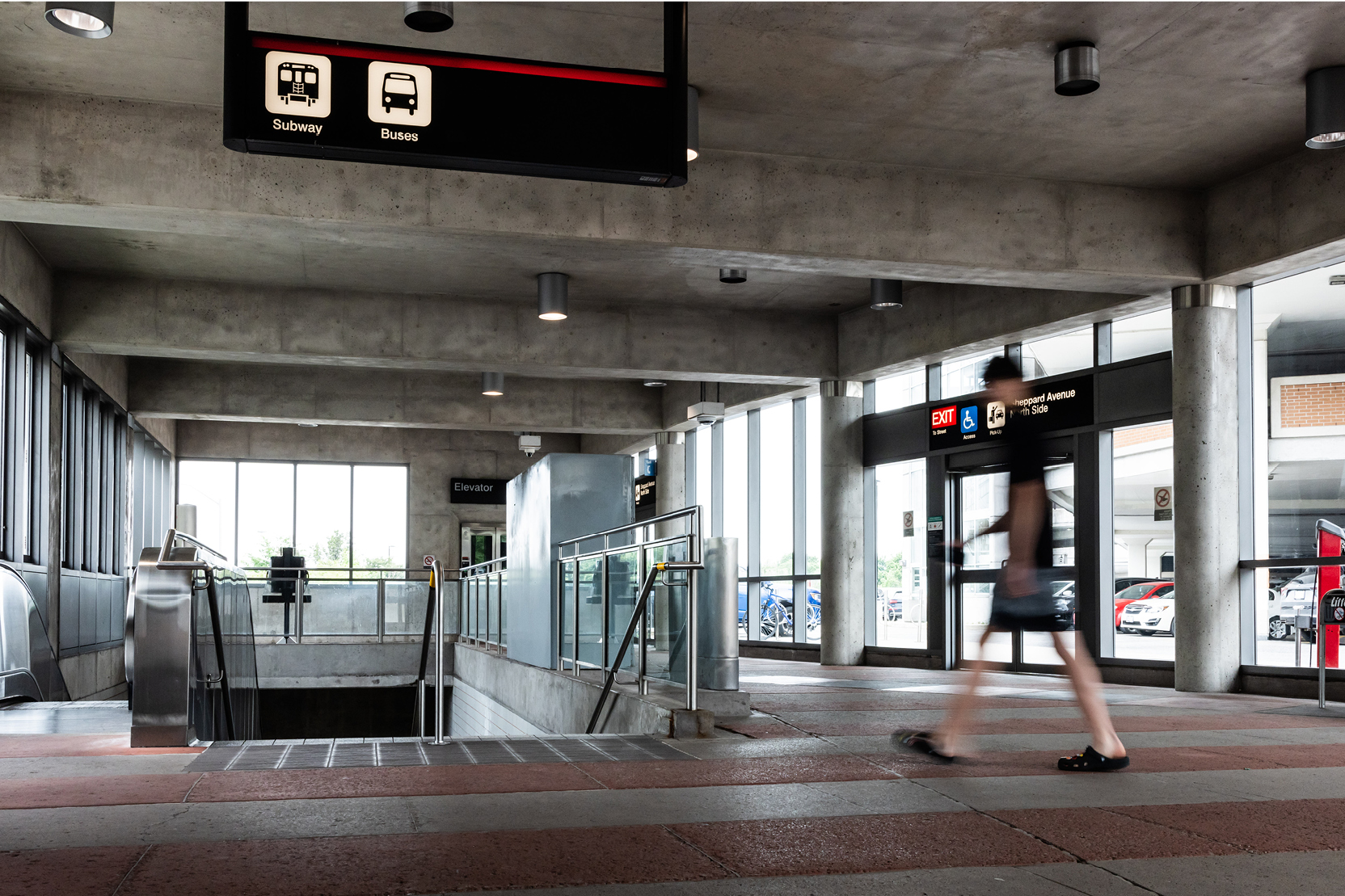 Transit station concourse with exposed concrete architecture, subway and bus wayfinding signage, an escalator to the platform, and an exit to Sheppard Avenue.
