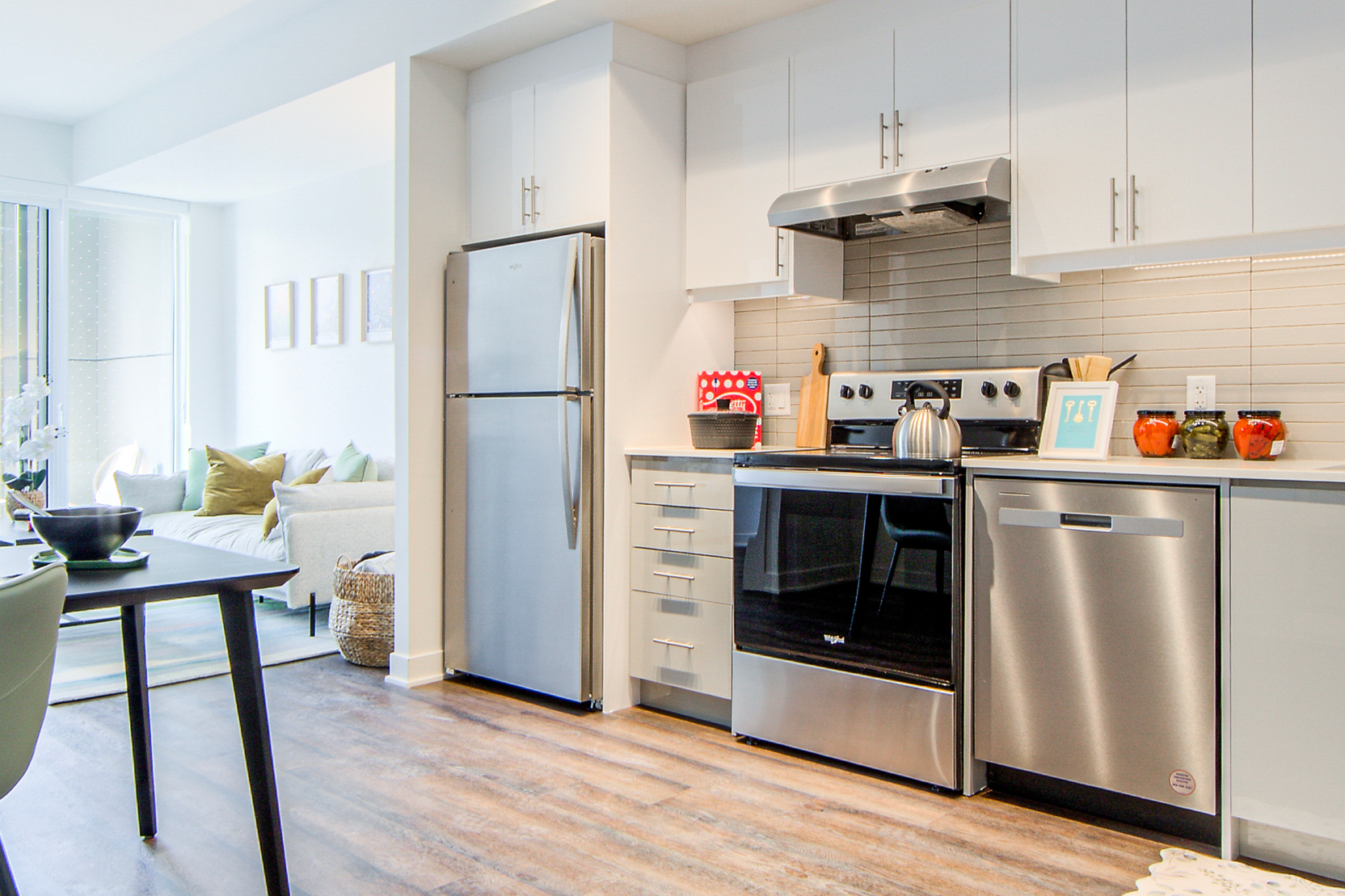 Modern open-concept kitchen with stainless steel appliances, white and gray cabinetry, linear tile backsplash, and wood-look flooring opening to a bright living area.