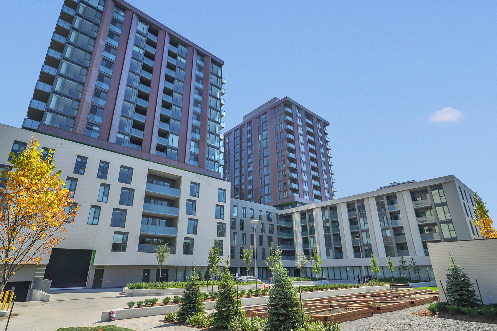 Exterior view of 3030 Pharmacy Avenue showing two high-rise towers with burgundy and gray façades above lower podium buildings, surrounded by landscaped courtyards with young trees and garden beds.