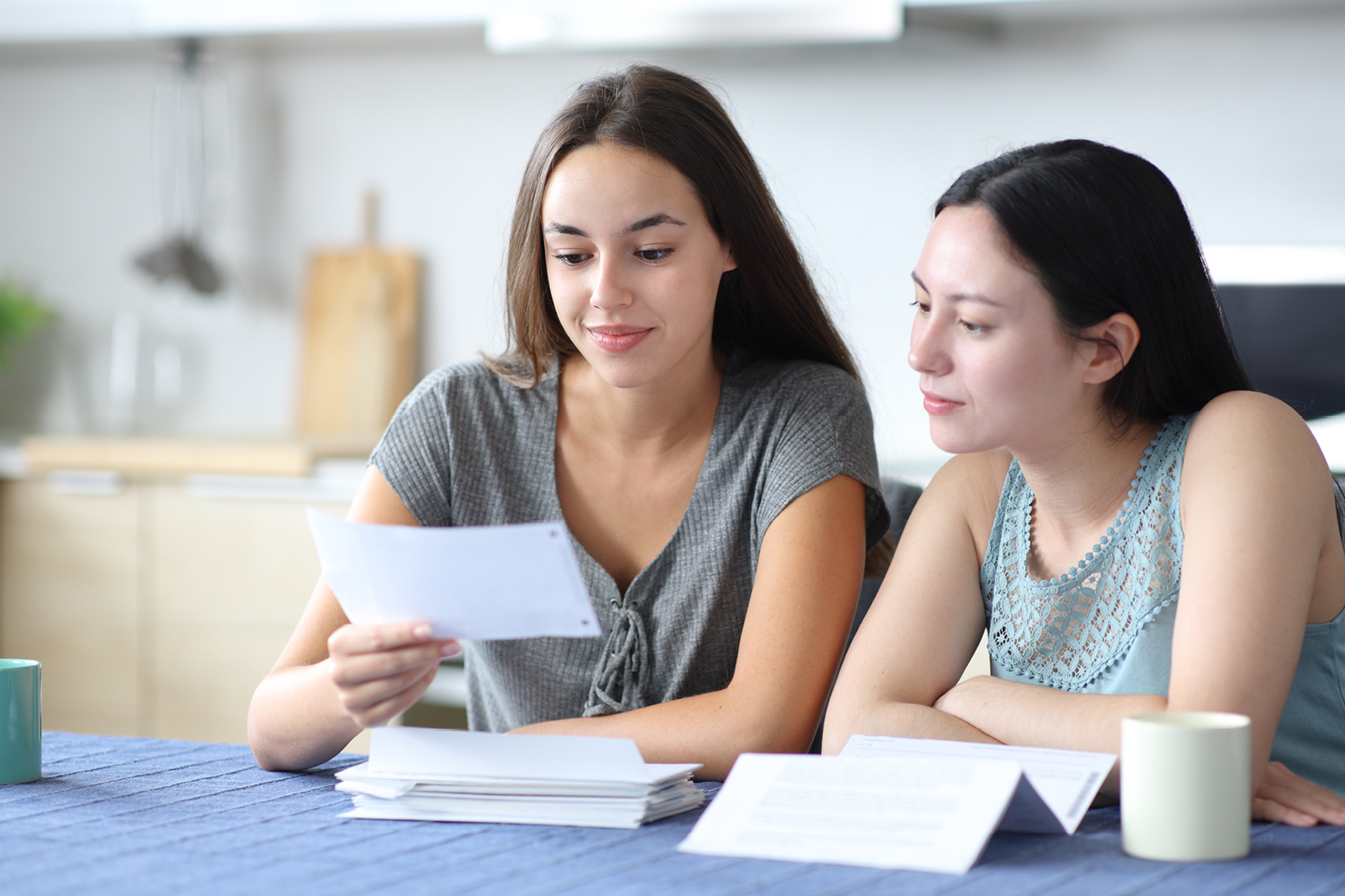 Two young women sitting together at a kitchen table, reviewing documents and bills over coffee.