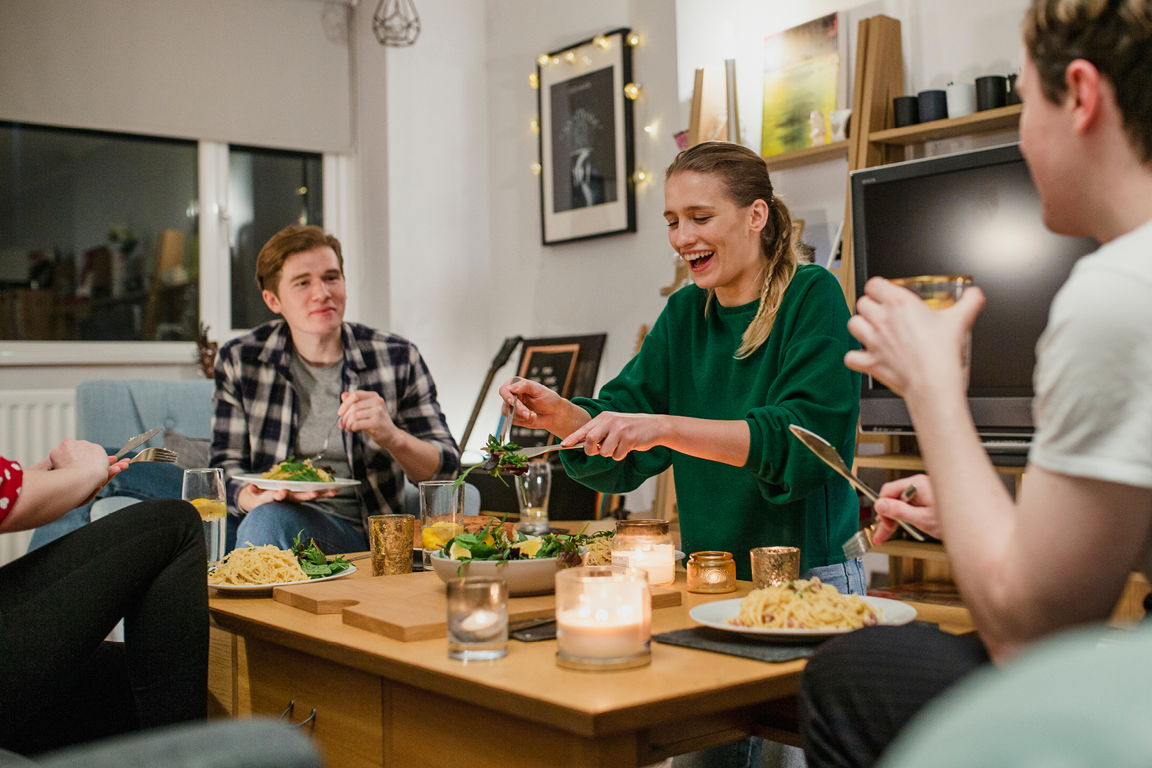 Friends laughing and sharing a meal together at a candlelit dining table in a cozy apartment living room.
