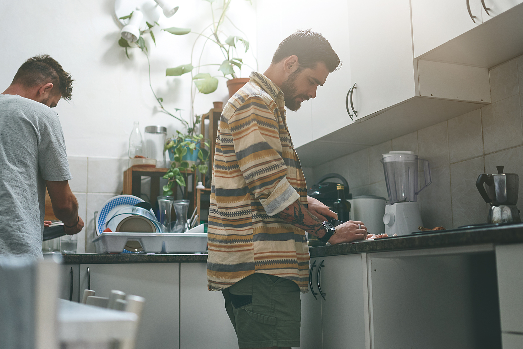 Two male roommates working side by side in a small kitchen, preparing food and washing dishes together.