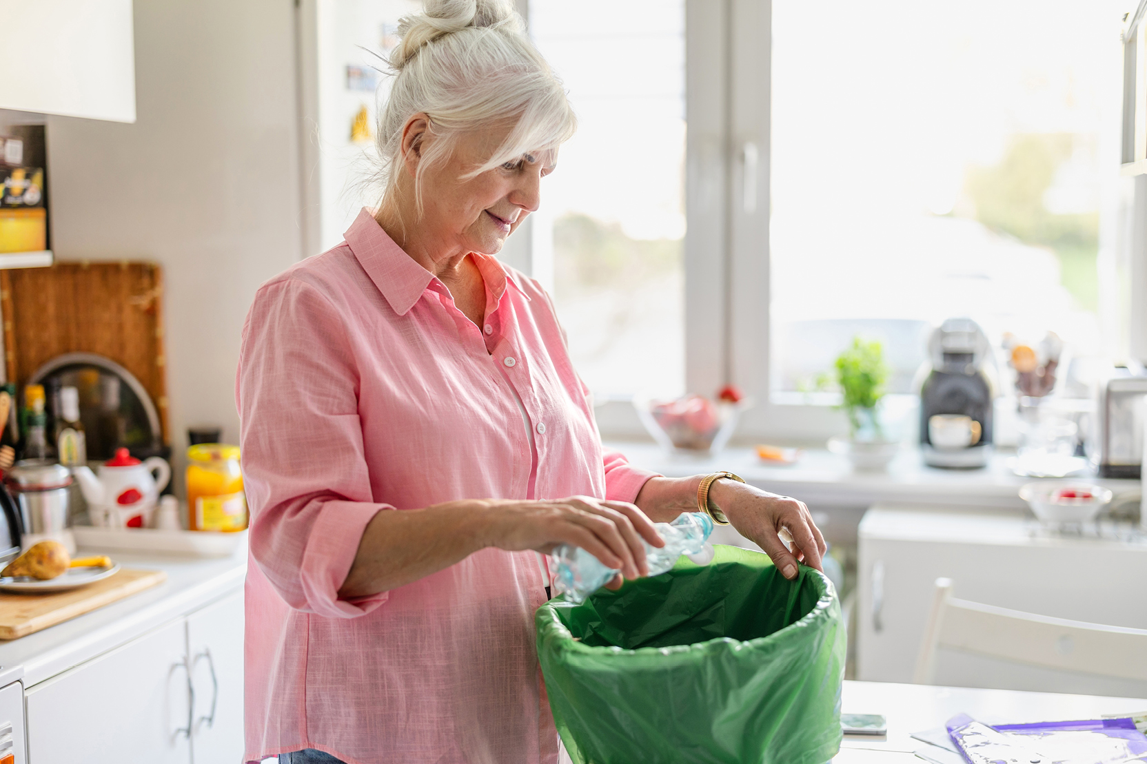 A woman places a plastic bottle into a green recycling bin in her kitchen.