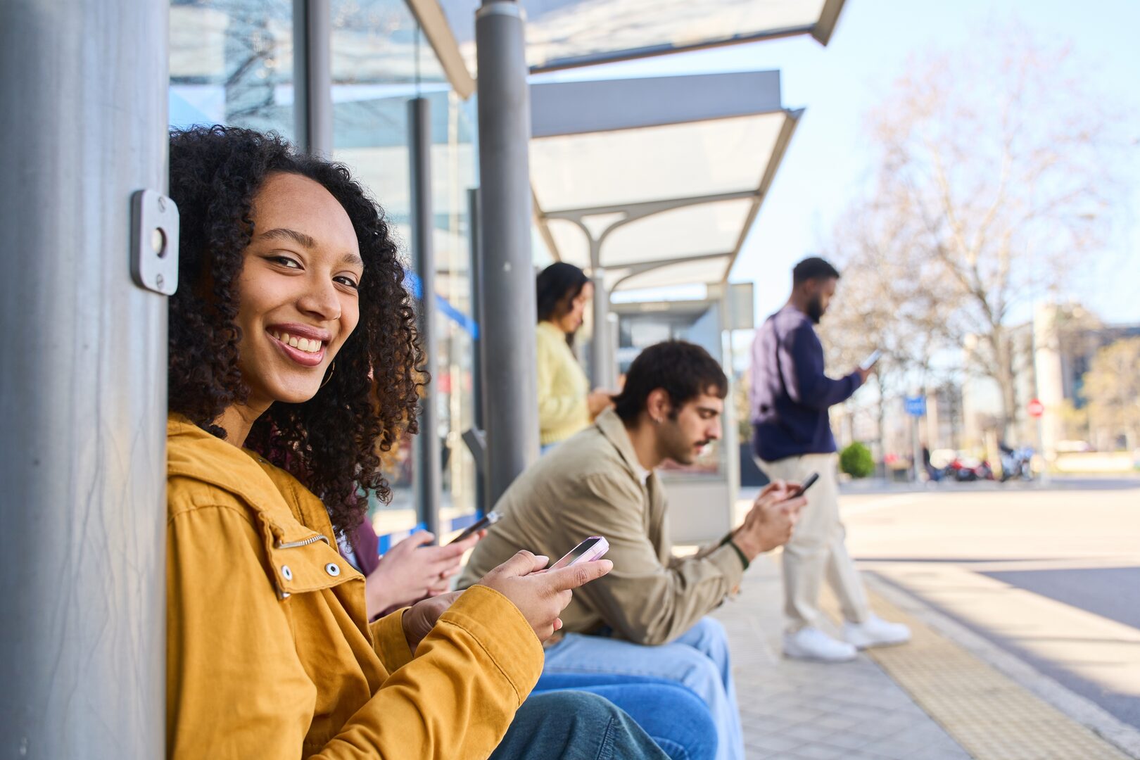 Smiling woman on her phone at a transit stop with other commuters in the background