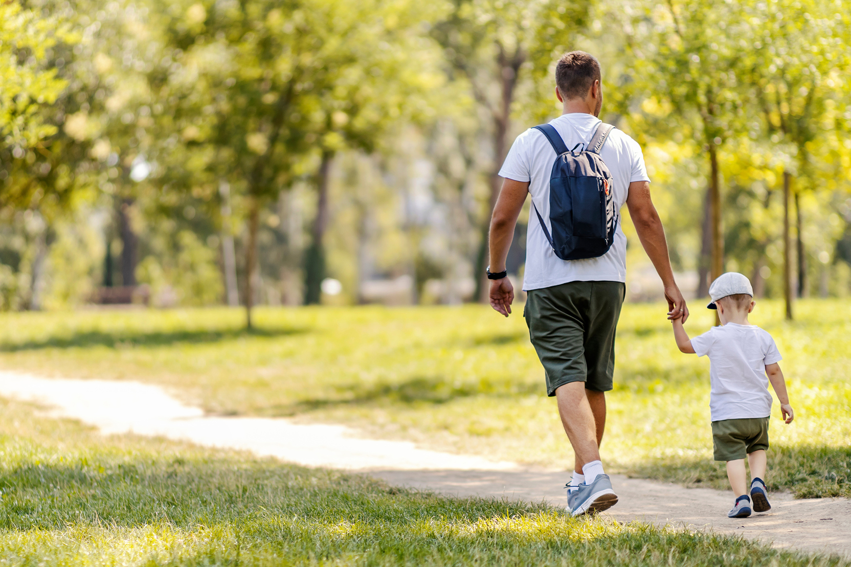 A father and young son walking hand-in-hand along a path in a park.