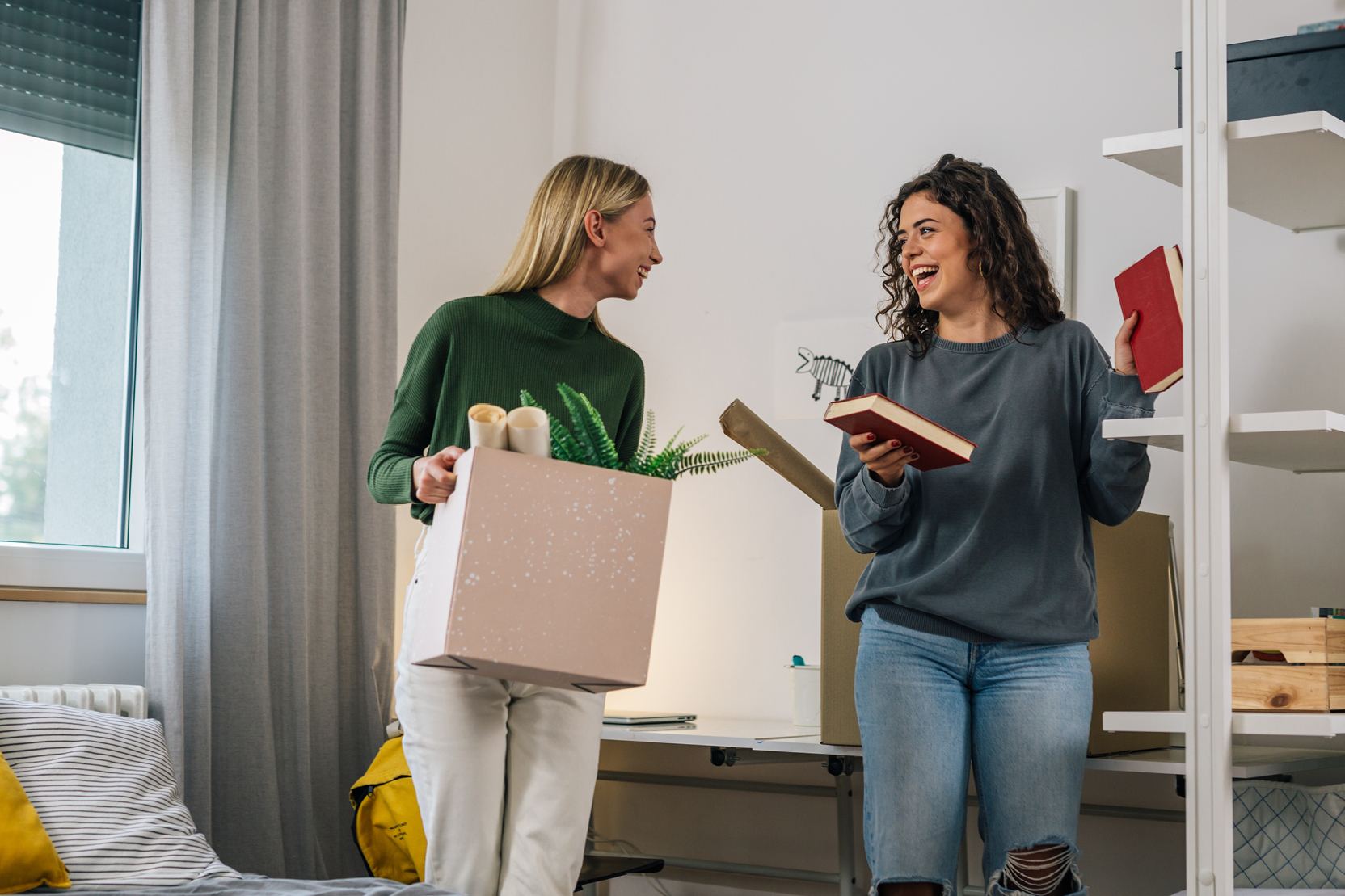 Two roommates laughing while unpacking moving boxes and books in a student apartment