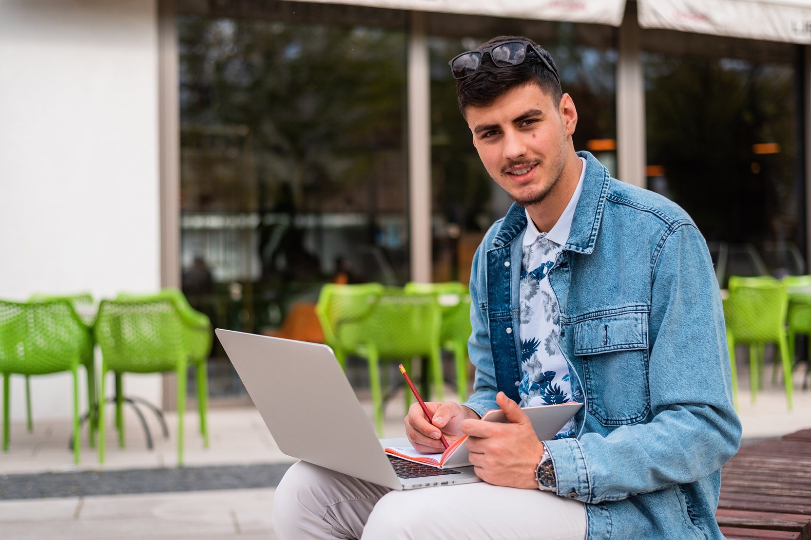 Student studying outdoors on a laptop with a notebook near a café patio