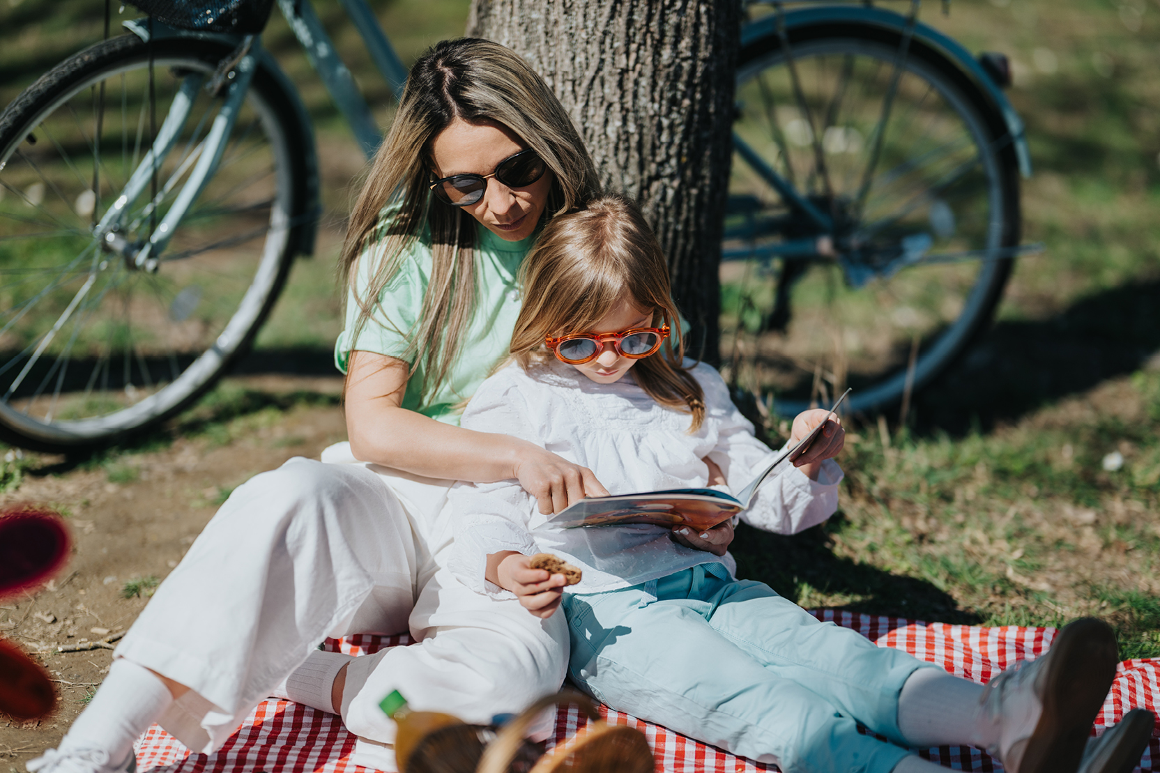 A mother and daughter lounge against a tree in a park to read a book.
