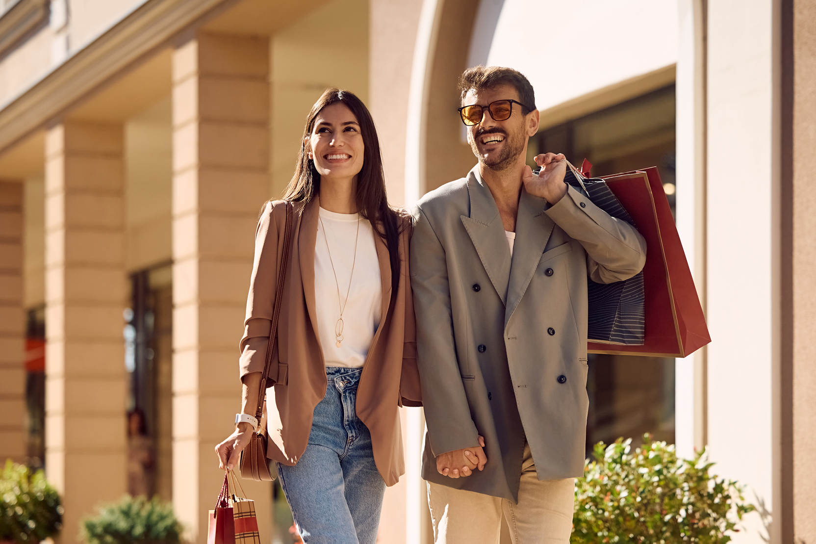 A couple walks along a sunny street holding hands and carrying bags after a shopping trip.