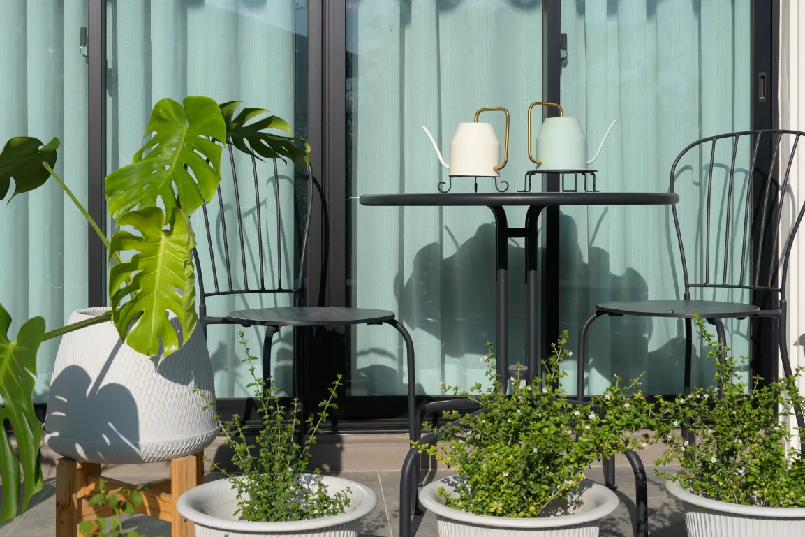 Apartment balcony with a small bistro table, two black metal chairs, and potted plants arranged in front of sliding glass doors.