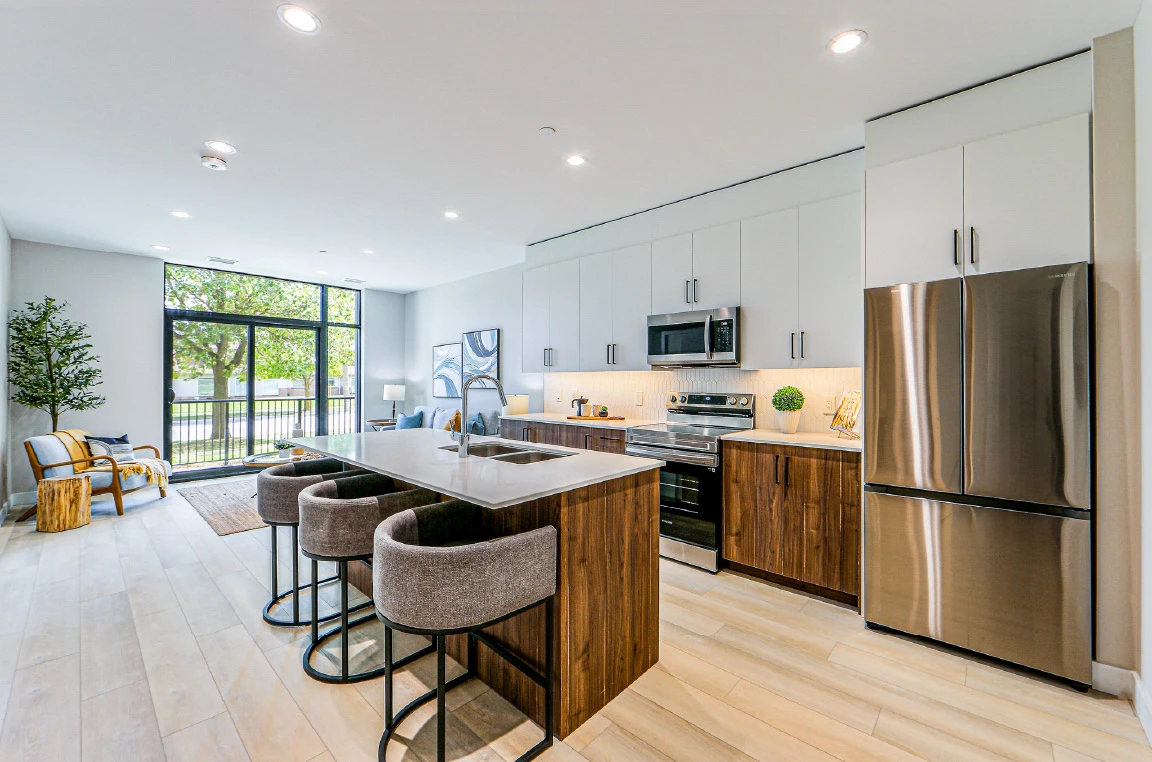 Modern open-concept apartment kitchen and living area featuring a large island with seating, white and wood-tone cabinetry, stainless steel appliances, floor-to-ceiling windows, and abundant natural light.