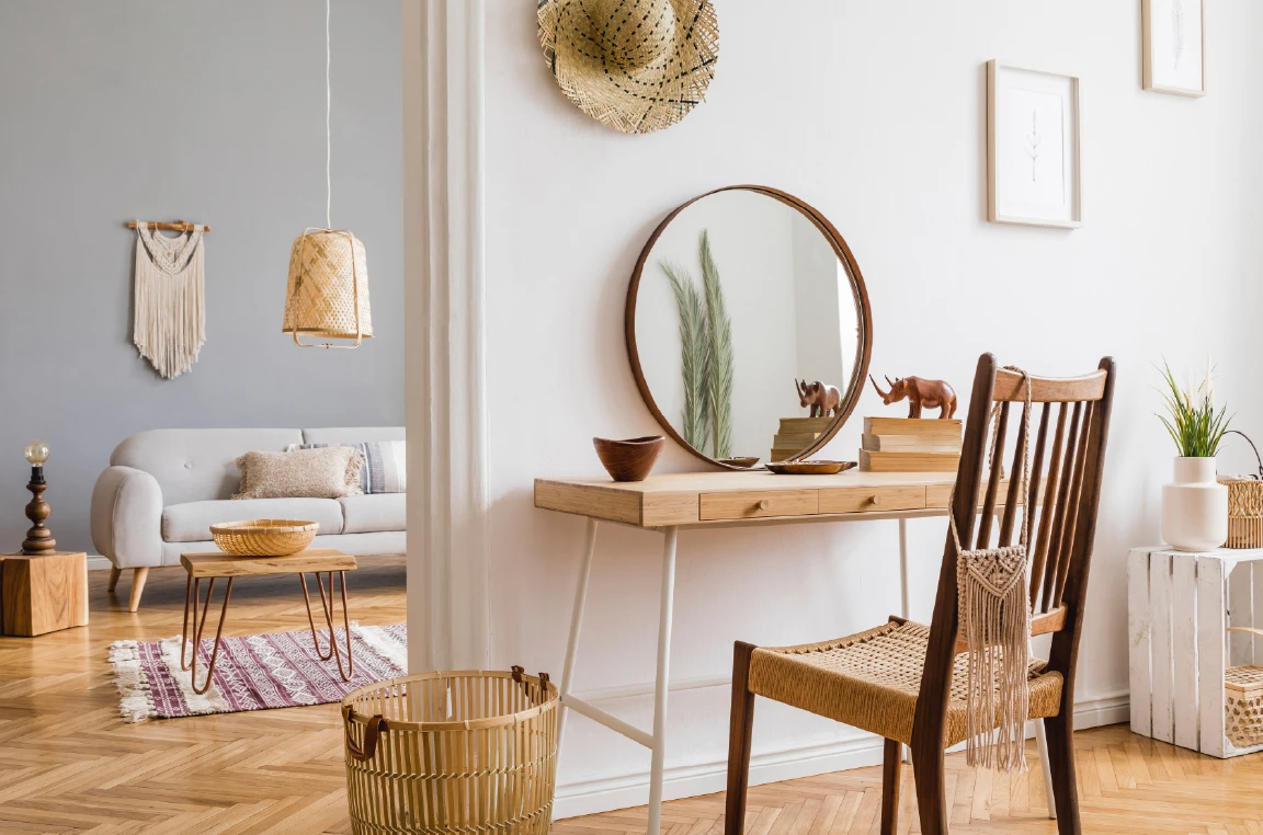 Bright apartment corner featuring a wooden vanity table with a round mirror and woven chair, complemented by neutral decor, indoor plants, and a cozy living area with a sofa, coffee table, and woven wall accents.