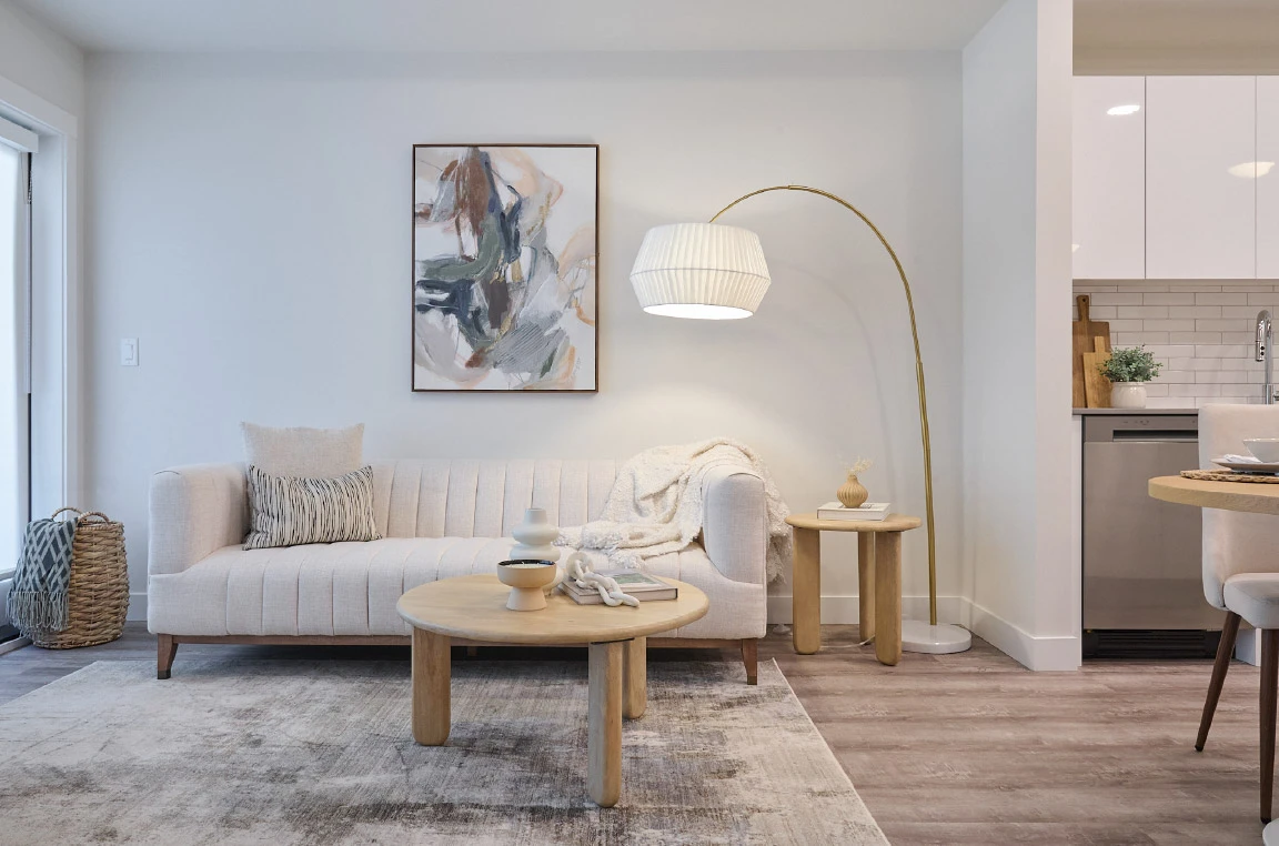 Modern apartment living room featuring a light-coloured sofa, round wood coffee table, floor lamp, abstract wall art, and an open-concept layout leading to a contemporary kitchen with white cabinetry.