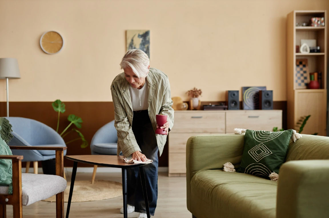 Person cleaning a coffee table in a modern apartment living room featuring a green sofa, armchairs, wood furniture, indoor plants, and contemporary decor.