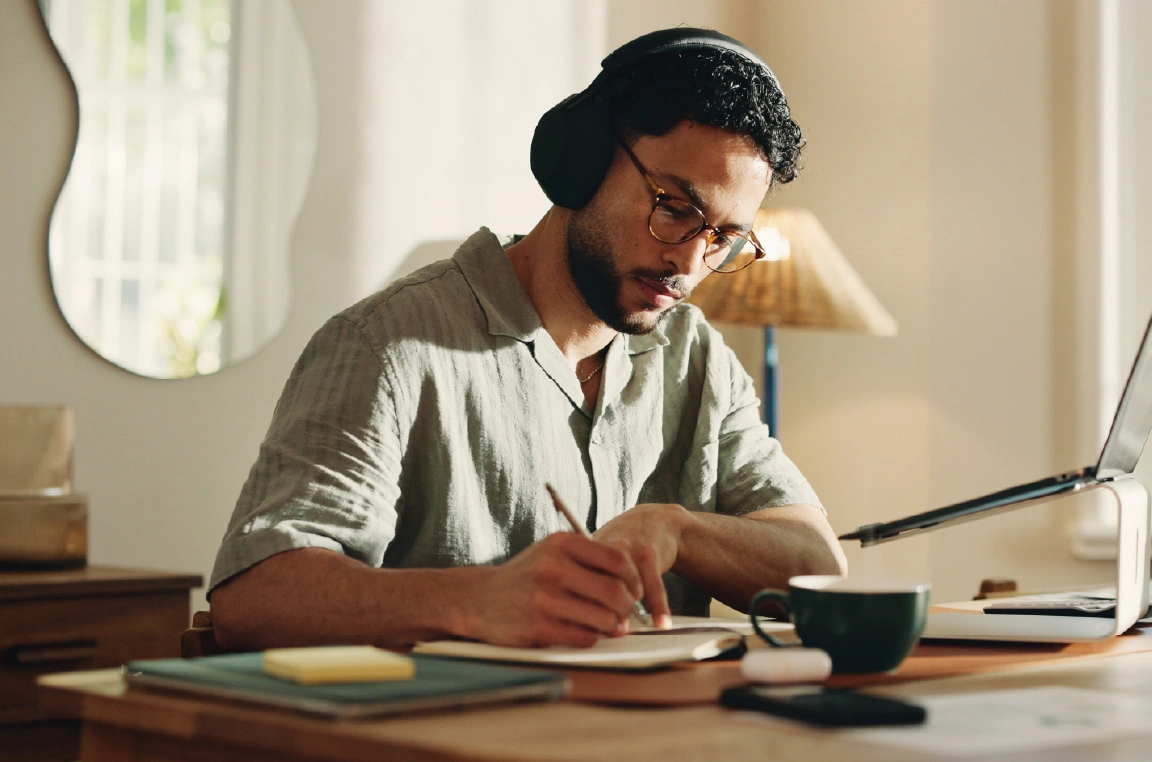 A man wearing headphones sits at a wooden desk in a bright home workspace, writing in a notebook beside a laptop on a stand. A coffee mug, sticky notes, and notebooks are arranged on the desk, with natural light streaming through nearby windows.