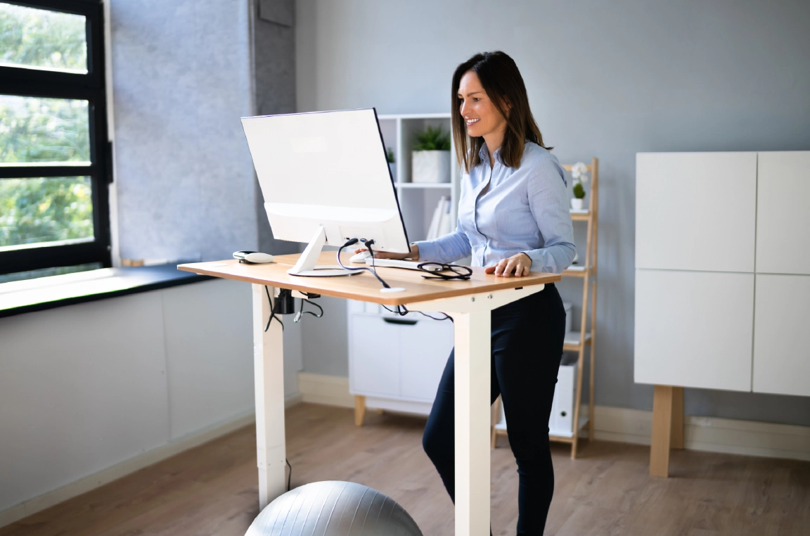 A woman with a calm smile works at a wooden standing desk with a yoga ball underneath. On the desk are her desktop computer, glasses, keyboard, and mouse. In the background, a large widow lets in natural light beside a shelf filled with plants.