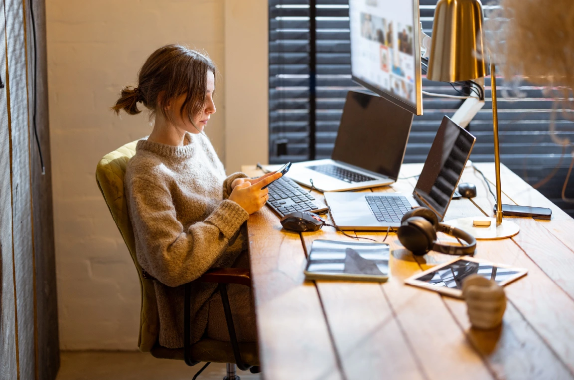 A young woman sits at a wooden desk, working on her phone while her laptop and desktop computer are open in front of her. On the desk are a plant, a lamp, an iPad, and headphones. She sits comfortably as she works. In the background, a wood plank wall and a window with closed blinds create a warm, enclosed atmosphere.