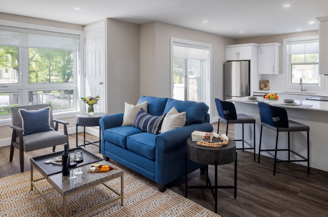Aura Apartments suite furnished with a two-seater blue couch, side chair, coffee table, and kitchen with an island and bright, open windows.